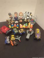 Full view of the entire toy lot showing bobbleheads, toy vehicles, action figures and assorted small toys arranged on a black surface.