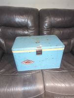 Vintage robin egg blue Coleman cooler on a brown couch showing full unit with latch and worn paint finish.
