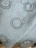 Close-up of pale muted teal linen fabric showing raised embroidered circular patterns in darker teal, cream, and chocolate thread.