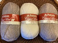 Three balls of Patons Classic Wool Worsted yarn, two pale tea colored and one cream colored, placed on a woven surface.