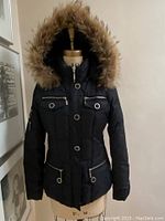 Black down-filled winter coat on a dress form showing full front with zippered pockets, silver buttons, and large hood trimmed with coyote fur.