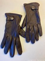 Both gloves laid flat, showing smooth dark chocolate leather with five fingers and snap button wrist detail.
