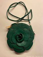 Full view of small green flower-shaped leather bag with long strap coiled above it.