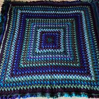 Full view of square blanket showing concentric square stripes in turquoise, sea blue, purple, and dark green with ruffled edges.