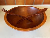 Large wooden salad bowl with two crossed wooden salad servers (fork and spoon) inside the bowl on a white surface.