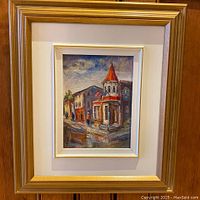 Framed small oil painting showcasing a street corner with a building featuring a pointed red roof and two flags.