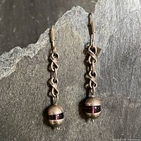 Pair of drop earrings showing amethyst spherical stones encased in half-circle sterling silver at top and bottom, connected by thick S-shaped chain links, laid flat against a dark textured background.