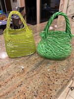 Two hand blown art glass baskets displayed on a countertop. One basket is bright yellow with horizontal stripes and the other is green with green swirl patterns. Both have twisted glass handles and are approximately 10 inches tall.