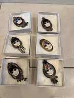 Six artist made brooches pinned in white gift boxes on floor, showing ceramic faces with metal frames and embellishments.