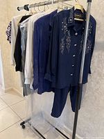 Full rack shot showing seven pieces of vintage women's clothing including pantsuits, blazers, and skirt sets mostly in dark colors and white, some with embellishments.