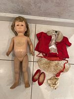 Photo showing the front of nude Chatty Cathy doll with blonde hair and the included clothing and shoes: red coat with fur collar, lace skirt, red shoes, socks.