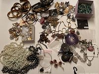 Overhead photo showing the full assortment of jewelry items including pearl necklaces, bracelets, chains, brooches, and watches arranged on a white surface.