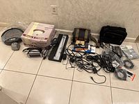 Photo of miscellaneous electronics including round ceiling lamp in box, black and white desk lamp, older Roomba vacuum, clock radio, fluorescent lamp, cables, and electronic accessories on tiled floor.