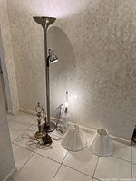 Four lamps and two white lampshades arranged on floor near wall on tiled floor. Floor lamp is tall with metal finish, smaller table lamps show metal wear, glass table lamp has clear glass body.