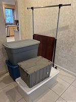 Stacked plastic totes with lids and folded Samsonite card table leaning against wall with metal clothing rack behind