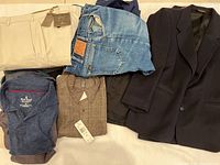 Assorted men's clothing displayed including beige trousers, denim jeans, brown trousers with tag, navy and dark blazer, and button-down shirt.