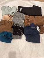 Assorted clothing items including pants, sweaters, a bra, and scarf laid out on a light surface.