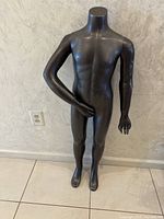 Full frontal image of a black matte mannequin representing an older child size, standing on white tiled floor against a light textured wall.