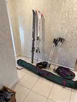 Two pairs of skis standing against wall with two sets of ski poles and three ski bags on ground