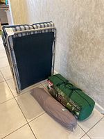 Portable folding cot with blue and beige striped fabric, standing upright next to two tents in their bags on the floor.