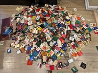 Large pile of hundreds of matchbooks with a few postcards mixed in, showing various colors and designs scattered on floor.