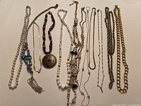 Photo showing 12 necklaces laid out, including faux pearl strands, bead necklaces, chains, and pendant necklace with bronze round charm.