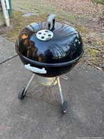 Full view of Weber round kettle-style charcoal grill with lid on concrete patio outdoors. Shows black grill body, metal legs and wheels, and ash catcher underneath.