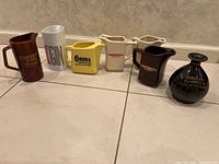 Photo showing seven assorted pitchers and one old bottle arranged in a row on tiled floor.