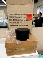 Black round gift box sitting on a cardboard box with a plastic wrapper partially around the box.