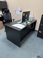 Full view of the black desk showing its shape and clutter on top, including a computer monitor and keyboard.