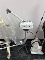 Full view of the facial steamer on black tripod stand with caster wheels, the main unit covered partially in plastic and accompanied by power cable.