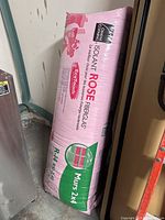 Photo of an unopened pink bag of Owens Corning Isoflam Rose fiberglass insulation leaning against a wall, detailed labeling visible, shows product for 2x4 walls, R14 value