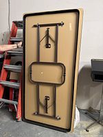 Underside of a wooden folding table showing beige laminate top and brown metal folding legs with locking mechanisms.