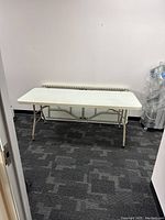 Full view of standard white folding table in rectangular shape against a neutral wall and patterned carpeted floor.