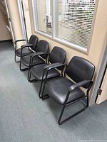 Four black waiting room chairs lined up in an office setting next to a glass-paneled wall, showing padded seats and backs with black metal sled frames and armrests.