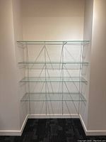 Five clear glass shelves mounted on white metal support rails affixed to the wall forming a corner shelf unit.