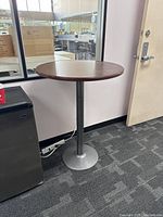 Round high top table shown beside wall and a mini fridge, highlighting wood veneer top and metal pedestal base.
