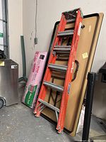 Orange Louisville fiberglass ladder leaning against folding table in storage area.