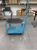 Front angle view of the blue metal rolling buggie showing two shelves and handle on a warehouse floor.