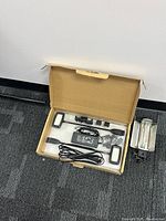 Open cardboard box showing two LED flood light arms with rectangular heads, power adapter and cables, and mounting attachments inside a clear plastic tray.