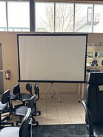 Front view of large white projector screen with black border, mounted on tripod stand in an indoor setting with office chairs nearby.