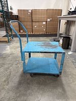 Side view of blue metal rolling cart showing two shelves, curved handle, and four wheels on concrete floor.