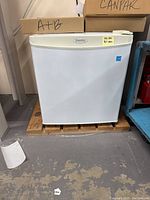 Front view of the small white Danfy fridge sitting on a wooden pallet with boxes on top.