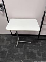 Full view of small white table showing rectangular white top and metal T-shaped base on dark carpet floor beside a wall.