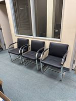 Three black and silver waiting room chairs arranged side by side against a beige wall under a window.