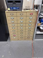 Front view of beige metal cabinet showing 24 small drawers and label holders