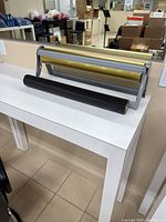 Full view of the wrapping paper cutter with the gold and black rolls on a white table in a room with boxes in the background.