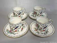 Four Aynsley demitasse cups and saucers displayed from above, clearly showing the intricate floral and bird pattern and gold rim.
