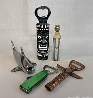 Lot of five collectible bottle openers and hourglass timer on white background. Includes Hawaii tiki face opener, shark opener, green handled opener, bronze totem pole opener with wings, and small hourglass timer.