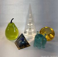 Five paperweights including a tall clear triangular prism, green pear, black pyramid, blue-green turtle, and yellow acrylic faceted round paperweight.